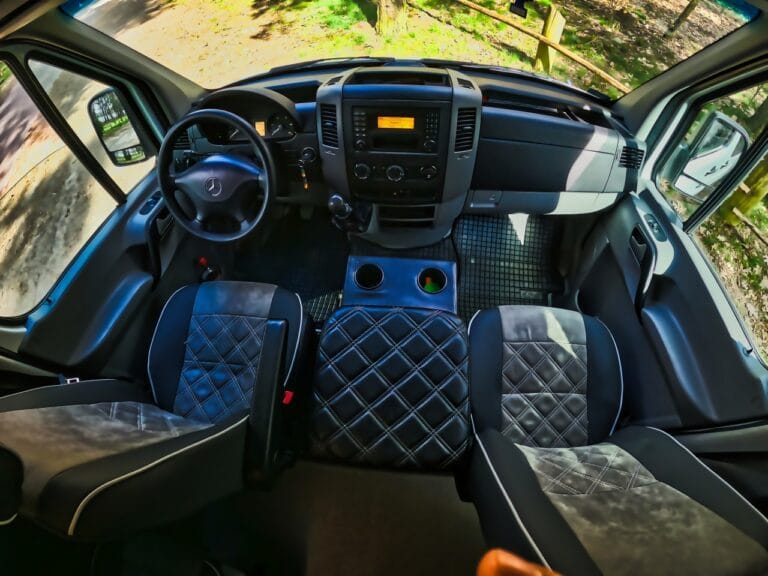 Widok wnętrza pojazdu, uwydatniający zajęty fotel kierowcy, deskę rozdzielczą i kierownicę z tapicerką ze wzorem w romby.