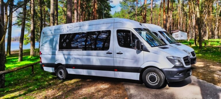 Biały bus zaparkowany na ruchliwej żwirowej drodze obok zalesionego obszaru ze zbiornikiem wodnym w tle.